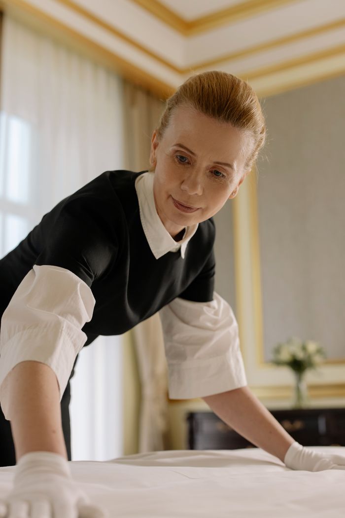 creative-03 A housekeeper in uniform skillfully arranges bed linen in a luxurious hotel room.