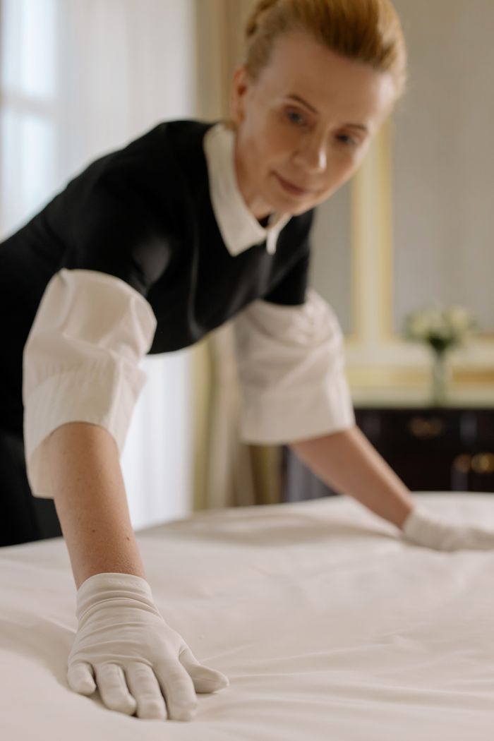 mobile-01 A housekeeper elegantly arranges bed sheets in a luxurious hotel room, showcasing hospitality expertise.