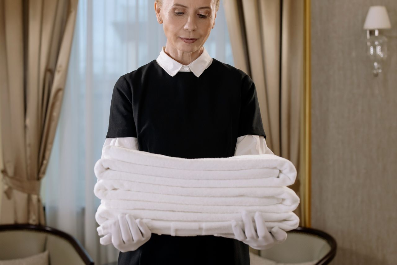 brand-01 Professional housekeeper holding a stack of fresh towels in a luxurious hotel room, showcasing elegance and service.